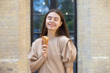 close-up of a smiling brunette girl with closed eyes eating ice cream in a waffle cup against a brick wall background clipart