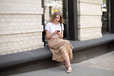 Güzel bir kadın, sarışın, akıllı telefon kullanıyor ve sokakta tuğla bir binanın yanında oturuyor.