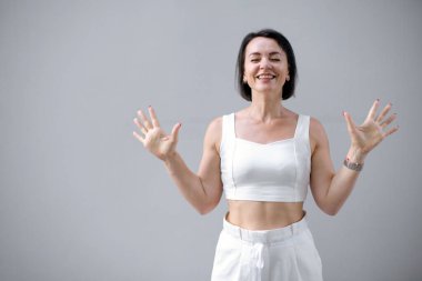 Beyaz takım elbiseli yakışıklı kadın kollarını gri yalıtılmış arka planda kaldırmaktan zevk alır. Beyaz kızın duygusal portresini kapat.