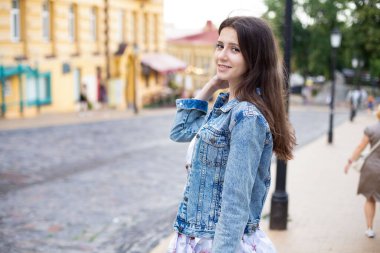 Kot ceketli güzel bir kız sokağın ortasında dururken kameraya bakıp gülümsüyor.