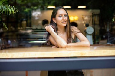 beautiful girl siting at the table in the inside of cafe on the background of neon sign. view from the street through the glass