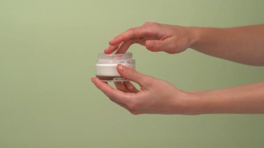 Bir kadın el, cam şeffaf bir model kavanozundan kremi alır ve onu parmağıyla tenine sürterek yeşil izole edilmiş bir arka plana sürer. Kişisel bakım için güzellik ritüelleri kavramı.