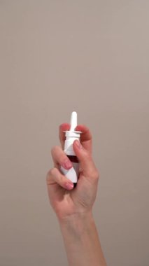 Dikey videoda kadın eli, cam bir burun spreyi kavanozunu tutuyor ve bej arka planda püskürtücüye bastırıyor. Soğuk algınlığı, virüs tedavisi, alerji mevsimi ve günlük konfor kavramı