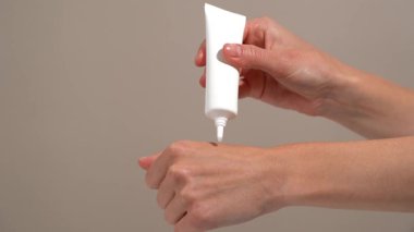Genç bir kadının eli beyaz model tüpten cilde serum uyguluyor ve bej arka planda parmaklarıyla ovuşturuyor. Özbakım, susuzluk ve ışıl ışıl cilt için güzellik ürünü kavramı