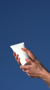 Beyaz model tüp tutan kadın eller, onu açıyor ve avuçlarının içine açık mavi gökyüzüne krem sürüyor. Doğal güzellik, hidrasyon ve yaşam tarzı cilt bakımı için günlük yüz kremi kavramı