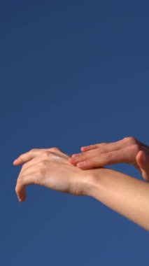 Kadın elinin dikey videosu açık mavi gökyüzüne sürttüğü bir krem lekesi. Sağlıklı cilt ve sağlık için organik kozmetik çözümler kullanma fikri.