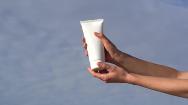 Kadın elleri beyaz losyon kremi tüpünü tutuyor ve bulutlu mavi gökyüzüne karşı avucunun içine sıkıştırıyor. Hidrasyon, vitamin ve cilt rutiniyle genç görünümü kavramı