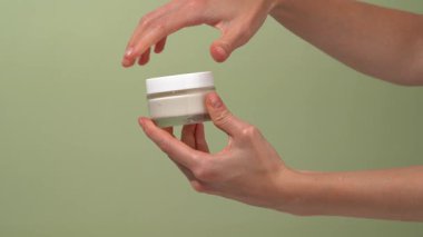 Dişi el kapağı açar ve cam şeffaf model kavanozundan kremayı parmağıyla alır ve yeşil izole arkaplandaki cilde kumaş lekesi uygular. Çevre dostu kozmetik çözümler kavramı