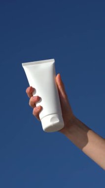 Açık mavi bir arka planda beyaz bir model tüpü tutan bir kadının dikey videosu. Yumuşaklık ve rahatlık için cilt temizleme ve nemlendirme kavramı