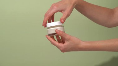 Bir kadın eli kapağı açar ve cam şeffaf bir taklit kavanozundan kremayı alır ve yeşil izole bir arka planda cilde uygular. Sağlıklı parıltı ve ışıl ışıl ten kavramı.