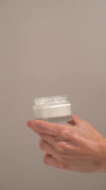 Bej arka planda şeffaf cam kavanozdan parmak kremi alan kadınların dikey videosu. Besleyici vücut kremi kavramı pürüzsüzlük, esneklik ve derin bakım sonuçları sağlar.