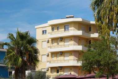 Kaldırımında sarı duvarlar ve birkaç palmiye ağacı olan modern Arnavut apartmanı. Gayrimenkul aramaları, kira listeleri, yabancı emlak alıcıları ve kıyı konutları için ideal.