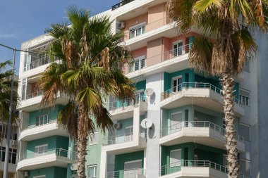 Modern balkon binası, palmiyelerle kaplı güneşli bir sahil şeridi boyunca duruyor. Yabancı emlak yatırımları, uzun vadeli kiralar ve Akdeniz konut piyasaları için mükemmel.