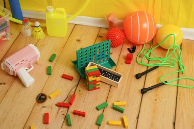 Yüksek açılı ahşap zemin ve çeşitli renkli oyuncaklar. Oyunların, topların ve blokların karışımı eğlenceli ve kaotik bir sahne yaratır. Parlak, canlı renkler.