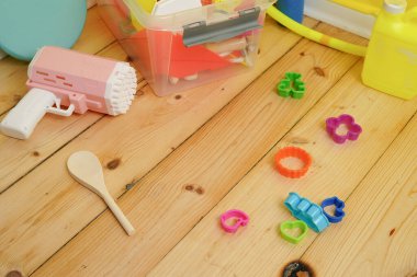 Açık ahşap zemine saçılmış renkli çocuk oyuncakları. Sahnede pembe bir balon tabancası, plastik hamur kesiciler, bir depolama kutusu, çocukluk eğlencesi yaratıcılığını çağrıştıran bir his var..