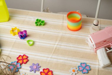 Bir çocuğun yaratıcı oyun seansına yüksek açılı bir bakış açısı. Gökkuşağı yayı gibi renkli oyuncaklar, pembe silah, ve keçeli çiçekler çizgili bir kumaşa saçılmış. Parlak, doğal ışık. Eğlenceli ve nostaljik.