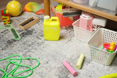 Bir çocuk için güvenli bir ortam değil. Zararlı kimyasallarla dolu sarı bir konteynır renkli oyuncaklar arasında dağınık bir halının üzerine bırakılıyor. Kazara ev halkını zehirleme riskini vurguluyor..