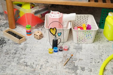 Bir çocuğun oyun odasının kaotik ama yaratıcı bir sahnesi. Renkli oyuncaklar, boyalar ve oyunlar gri bir halıya saçılmış durumda. Bu dağınık ortam aktif oyun, öğrenme ve çocukluk hayal gücünü akla getiriyor..