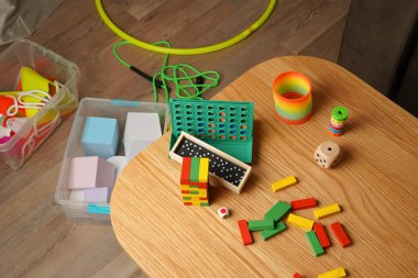 Klasik oyuncaklarla dolu yüksek açılı bir oyun odası manzarası ve ahşap bir masada oyunlar. Renkli bloklar, domino taşları ve zar evde eğlenceli, nostaljik bir aile eğlencesi sahnesi yaratır. Kapalı, doğal ışık.