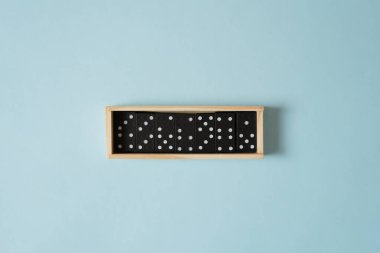Klasik domino oyunu setinin minimalist üst görüntüsü. Beyaz noktalı siyah fayanslar, sakin, pastel mavi bir arkaplan üzerine kurulmuş ahşap bir kutuda düzgünce düzenlenir. Temiz, basit, düz kompozisyon.