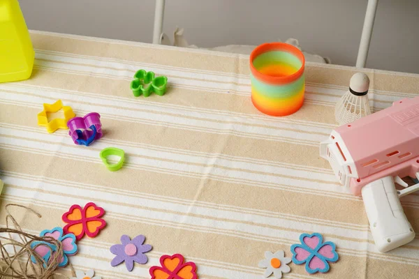 Bir çocuğun yaratıcı oyun seansına yüksek açılı bir bakış açısı. Gökkuşağı yayı gibi renkli oyuncaklar, pembe silah, ve keçeli çiçekler çizgili bir kumaşa saçılmış. Parlak, doğal ışık. Eğlenceli ve nostaljik.