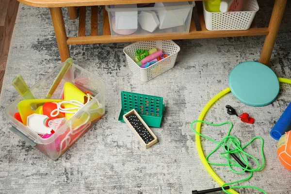 Dağınık bir oyun odası zemininin yüksek açılı görüntüsü. Renkli oyuncaklar, masa oyunları, spor aletleri gri bir halıya saçılmış, karmaşık bir oyun sahnesi yaratıyor. Çocukluk kavramı, oyun zamanı, düzensizlik.