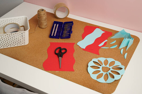 High angle view of a DIY crafting project. A cork board holds red and blue paper cutouts, scissors, and knives. This creative workspace evokes a feeling of hobby and handmade artistry. (188 chars)
