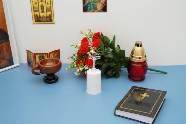 Dua için düzenlenmiş Hıristiyan dini eşyalar koleksiyonu. İçinde bir İncil, haç, ikonlar, kırmızı çiçekler, mavi bir masada beyaz bir duvara karşı duran bir mum var..