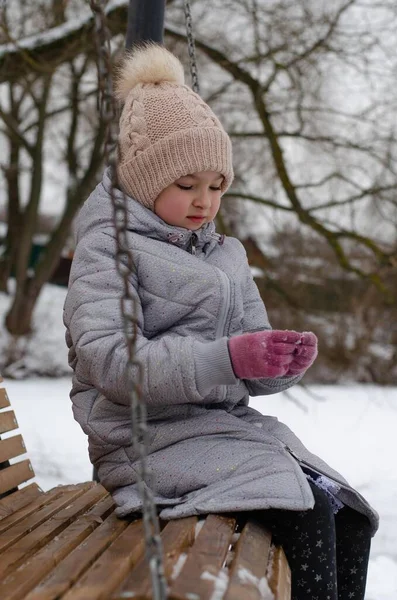 겨울 옷을 입고 겨울 공원에서 눈을 가지고 노는 모자를 쓰고 있는 소녀. 아이가 밖에서 나무 그네를 타고 노는 모습. — 스톡 사진
