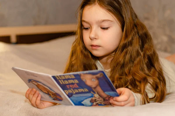 Ett barn, 7-årig flicka med långt blont hår läser en barnbok medan hon ligger på soffan. Bokeh ljus i förgrunden. Stockbild