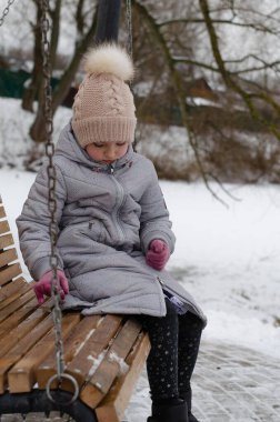 Kış kıyafetleri içinde bir kız ve kış parkında karla oynayan bir şapka. Çocuk dışarıda, salıncakta sallanıyor..