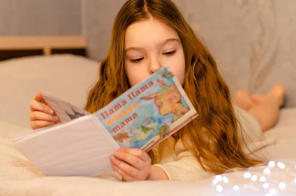 Ett barn, 7-årig flicka med långt blont hår läser en barnbok medan hon ligger på soffan. Bokeh ljus i förgrunden. Royaltyfria Stockfoton