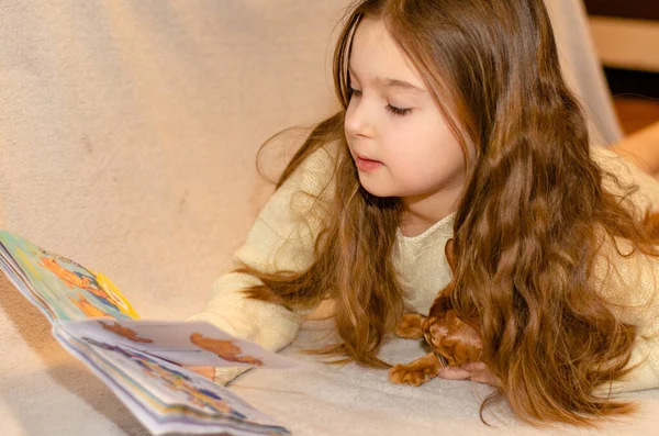 Un enfant de 7 ans avec de longs cheveux blonds lisant un livre pour enfants alors qu'il était allongé sur le canapé. Lumières Bokeh au premier plan. — Photo