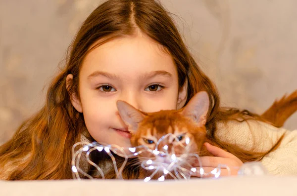 Una niña de 7 años con el pelo largo y rubio jugando con un gato en casa en el sofá. Concepto de mascotas y niños. — Foto de Stock