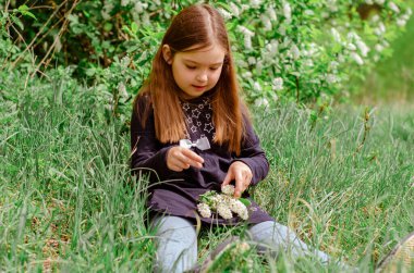 Uzun saçlı küçük bir kız çayırda oturmuş, kuş kirazının beyaz çiçeklerini kokluyor. Bahar çiçeği konsepti.