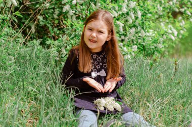 Uzun saçlı küçük bir kız çayırda oturmuş, kuş kirazının beyaz çiçeklerini kokluyor. Bahar çiçeği konsepti.