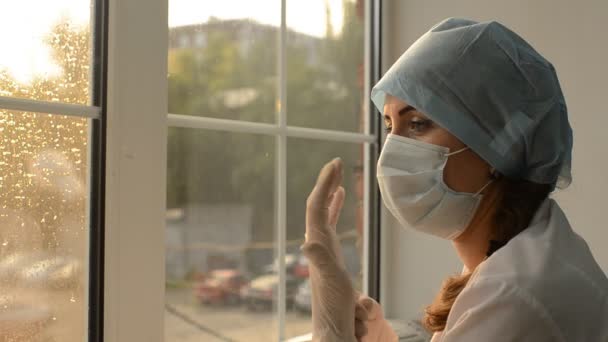 Ärztin in medizinischer Maske zieht Gummihandschuhe auf dem Fensterhintergrund an — Stockvideo