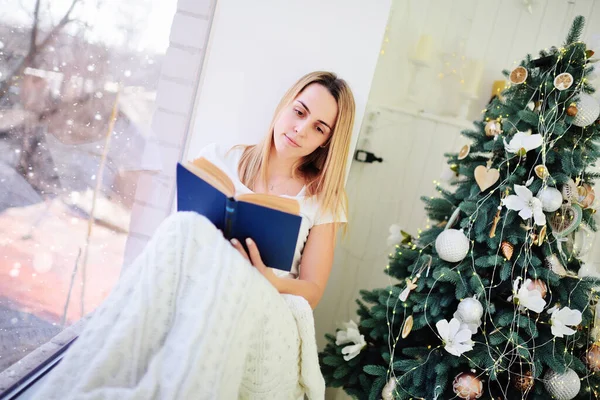 아름다운 여인이 창문 옆에 담요로 덮힌 책을 읽고 있어요 크리스마스 트리의 배경 옆에 앉아 있어요. — 스톡 사진