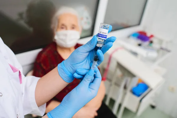 Uma enfermeira segura uma seringa e um frasco de vidro rotulado vacina covid-19 e se prepara para vacinar uma mulher idosa — Fotografia de Stock