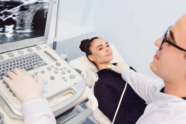 Lékař na moderní diagnostické klinice vyšetřuje štítnou žlázu pacienta mladé atraktivní ženy na ultrazvukovém přístroji — Stock fotografie