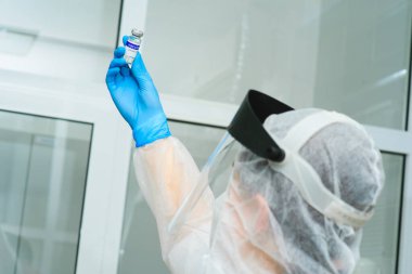 Doktor ya da koruyucu giysi, miğfer ve solunum cihazı giymiş bir PCR laboratuvar araştırmacısı üzerinde Covid-19 aşısı bulunan bir şişe tutuyor.
