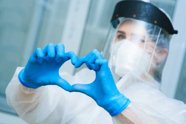 PCR laboratuarındaki araştırmacı koruyucu takım elbiseli, kasklı ve solunum cihazlı genç, güzel bir kadın, mavi lastik eldivenli bir kalp gösteriyor..