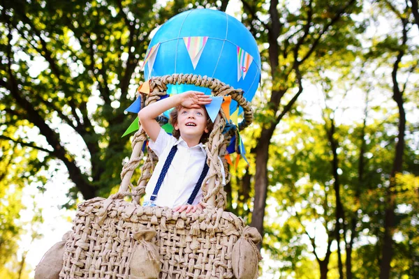 Menino com cabelo encaracolado em uma cesta de um balão azul sorri e olha para a distância contra o fundo da vegetação e do sol. — Fotografia de Stock
