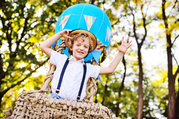 Маленький мальчик с вьющимися волосами в соломенной шляпе в корзине из воздушного шара улыбается и наслаждается путешествием. — стоковое фото