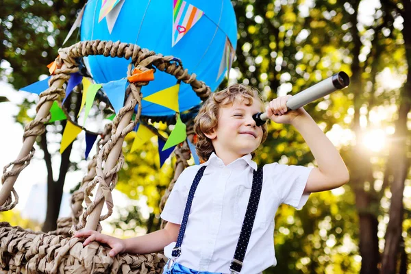 Kleine jongen met krullend haar met een telescoop in zijn handen tegen de achtergrond van een blauwe ballon mand glimlacht — Stockfoto