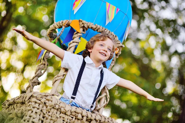 Small boy with curly hair in a basket of a blue balloon smiles and looks into the distance against the background of greenery and the sun. — Stock Photo, Image