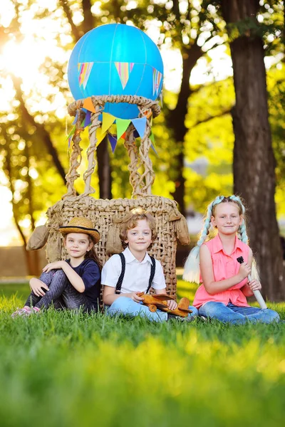 Üç küçük anaokulu çocuğu, arka planda bir sepet mavi balon ve güneş ışığına karşı çimlerin üzerinde otururlar.. — Stok fotoğraf