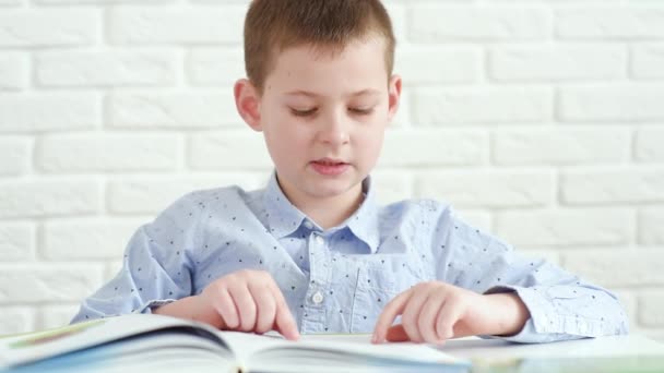 Uczeń dziecko czyta książkę siedzi przy białym stole na białym tle cegły — Wideo stockowe