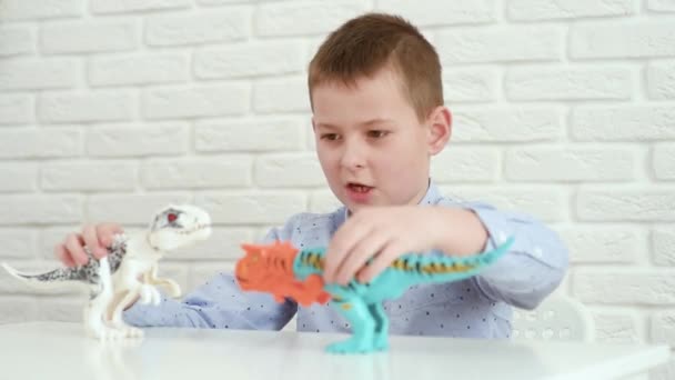 Niño sentado en una mesa blanca jugando una batalla de dinosaurios de juguete. — Vídeo de stock