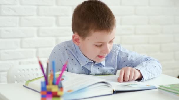 Uczeń dziecko czyta książkę siedzi przy białym stole na białym tle cegły — Wideo stockowe